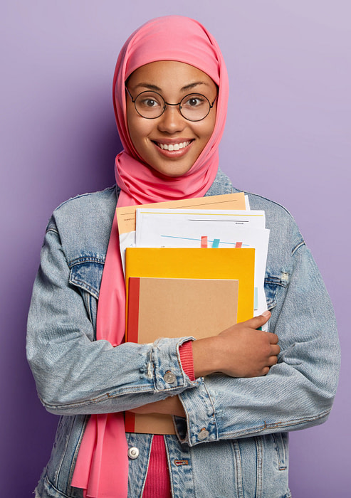 Beautiful young Arabian woman with dark skin, wears transparent spectacles, holds papers and notepad, has toothy smile, has religious views, studies at college, isolated against purple background